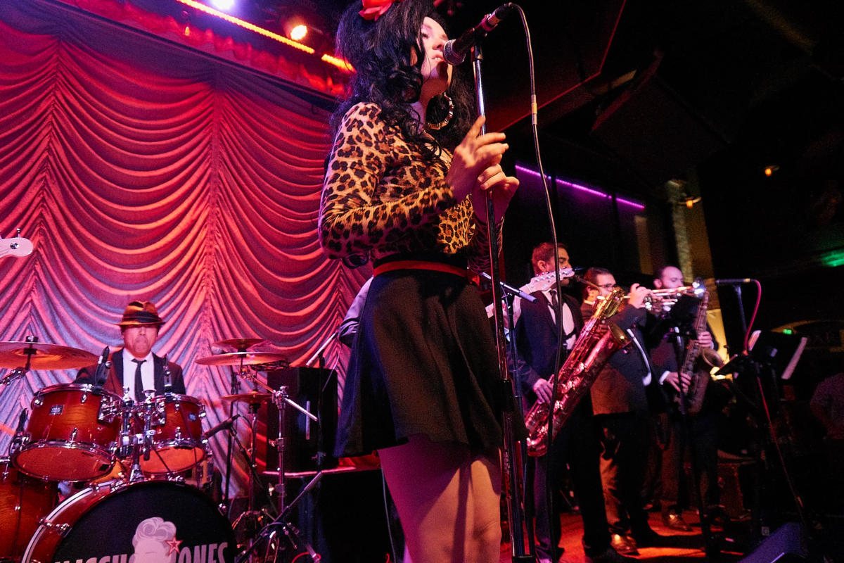 The full band on stage in red and pink light: the singer front and center, horns and drums behind